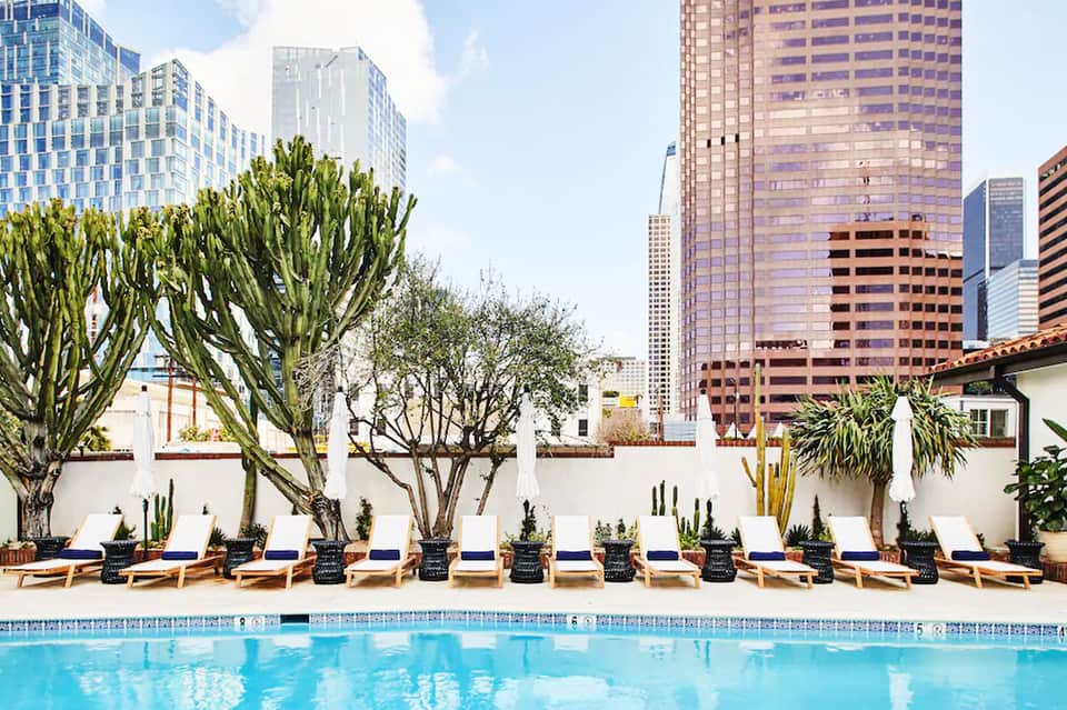 Modern hotel pool with lounge chairs, cacti, and white walls overlooking downtown city skyline