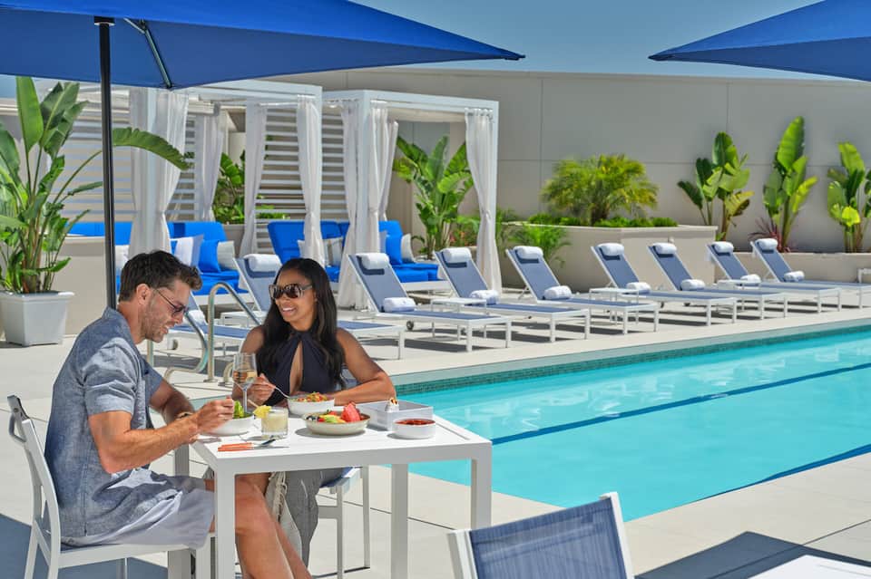 Couple dining poolside under blue umbrellas with lounge chairs and palm plants