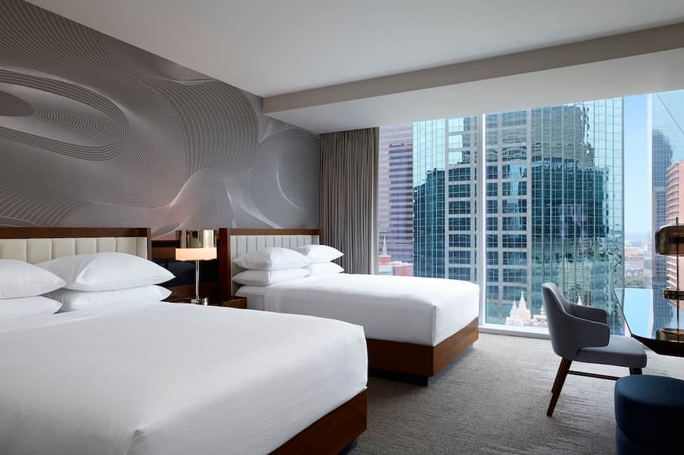 Modern hotel room with two queen beds, city skyline views through floor-to-ceiling windows, and abstract wall art