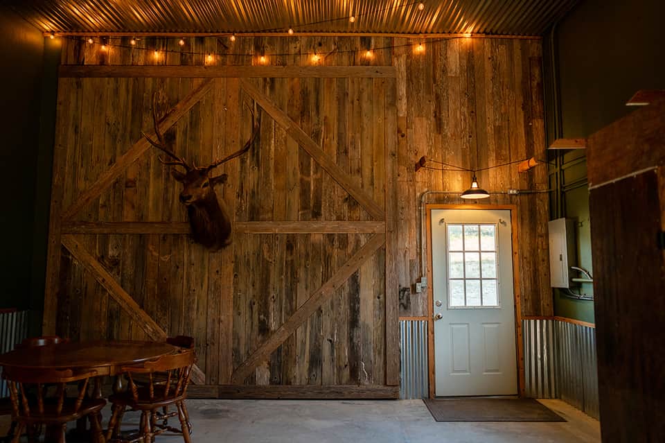 Rustic barn interior with wooden walls, exposed beams, deer mount, string lights, dining table, and white door