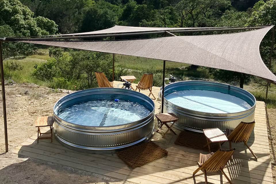 Two large circular hot tubs on wooden deck under shade sail with lounge chairs and forest backdrop
