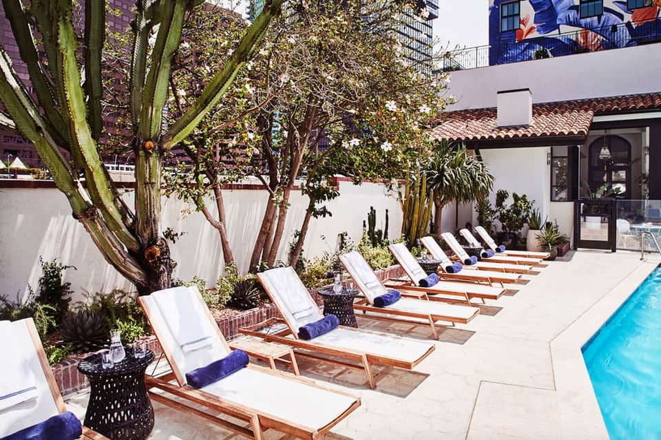 Poolside lounge area with wooden deck chairs, white cushions, and large trees providing shade