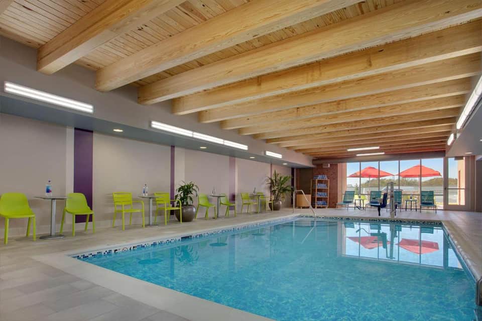 Indoor swimming pool with wooden beam ceiling, bright natural light, lime green chairs, and outdoor terrace view