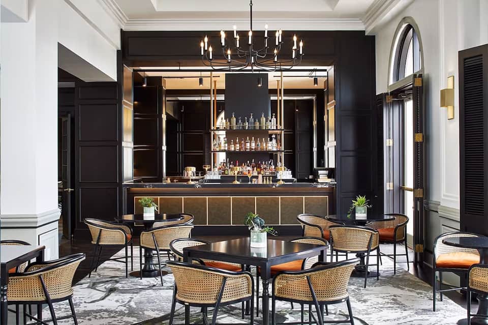 Upscale hotel bar lounge with black walls, illuminated bottle display, woven chairs, and modern pendant lighting