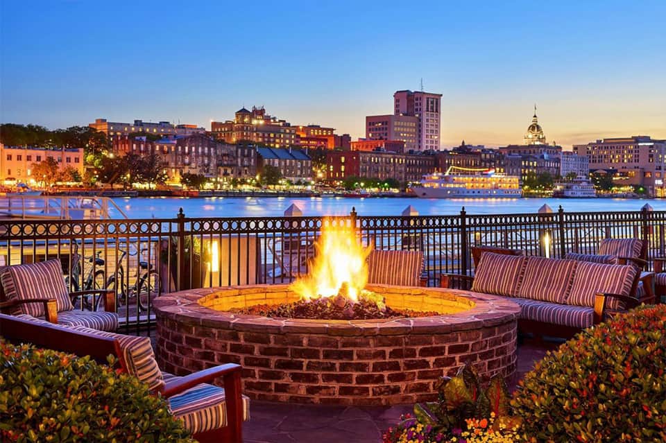 Waterfront fire pit lounge at dusk with city skyline, striped chairs, and brick railing