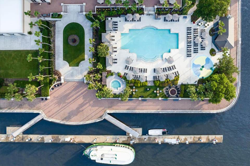 Aerial view of hotel pool complex with loungers, umbrellas, and waterfront boardwalk access