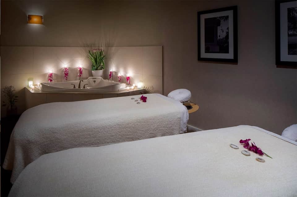 Spa treatment room with two massage beds, jacuzzi tub, and ambient candle lighting