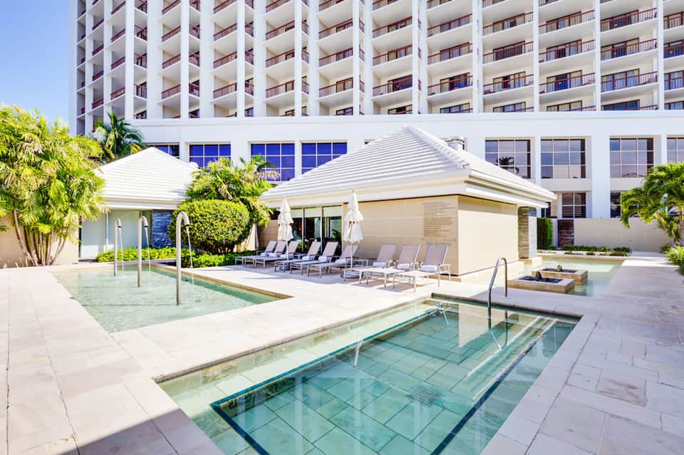 Resort pool deck with cabanas, loungers, white tent structures, and high-rise hotel building in background