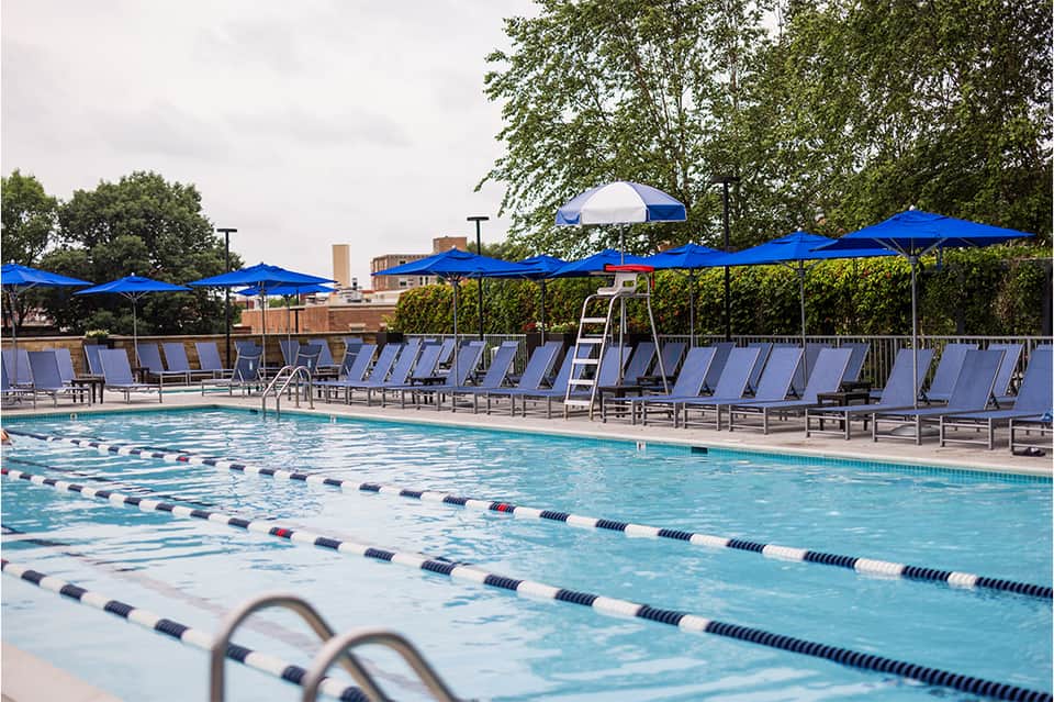 Lap pool with lane dividers, lounge chairs under blue umbrellas, and shaded seating area with trees.