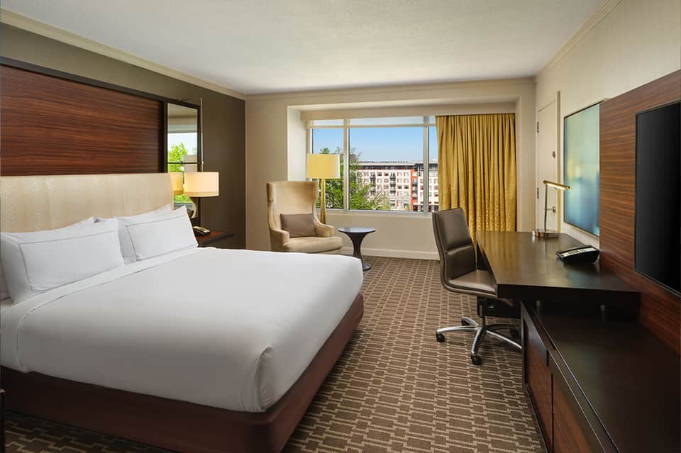 Hotel bedroom with queen bed, desk, chair, and city view from window