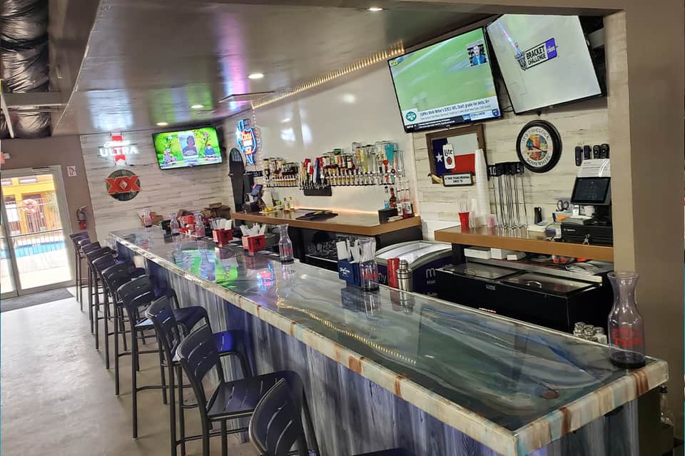 Sports bar interior with long counter, bar seating, TV screens, and shelving with bottles and sports memorabilia