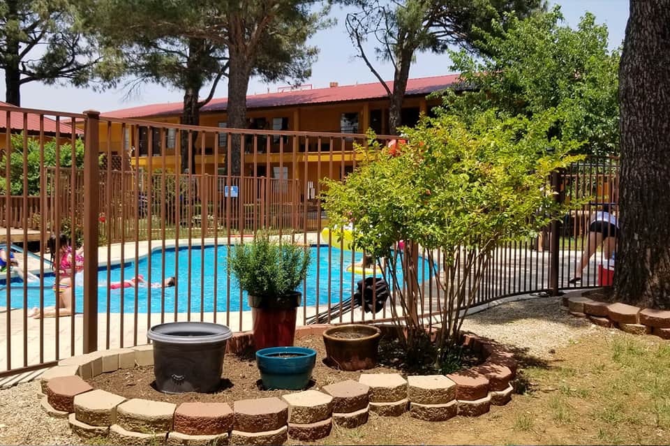 Fenced swimming pool surrounded by trees with brown building, fire pit area, and natural landscaping
