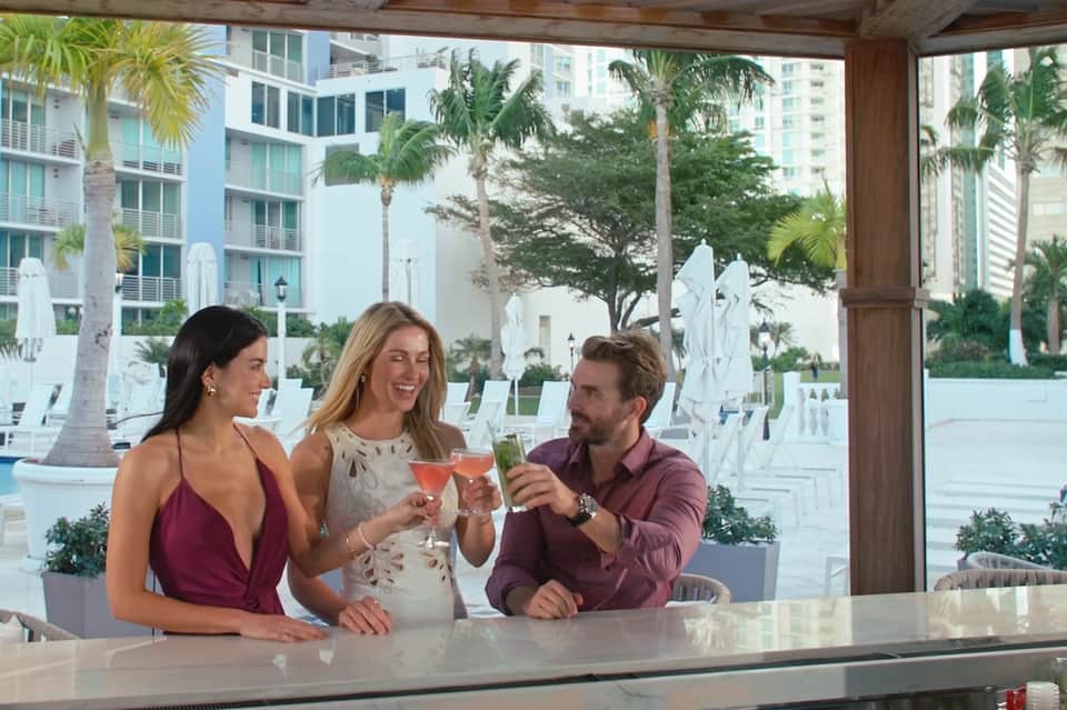 Three guests toasting cocktails at an indoor bar with tropical poolside view and palm trees