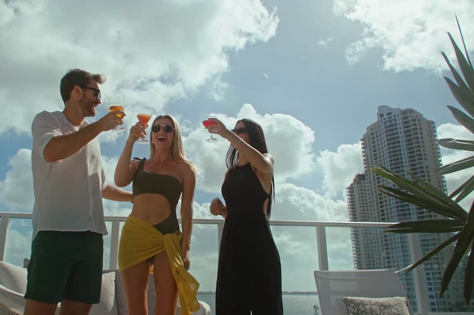 Three people toasting cocktails on a high-rise balcony overlooking city buildings and waterfront