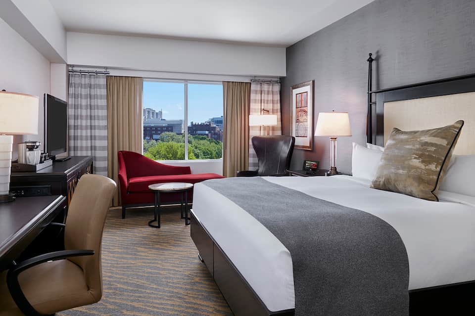 Modern hotel bedroom with king bed, city views from large window, burgundy accent chair, and contemporary furnishings