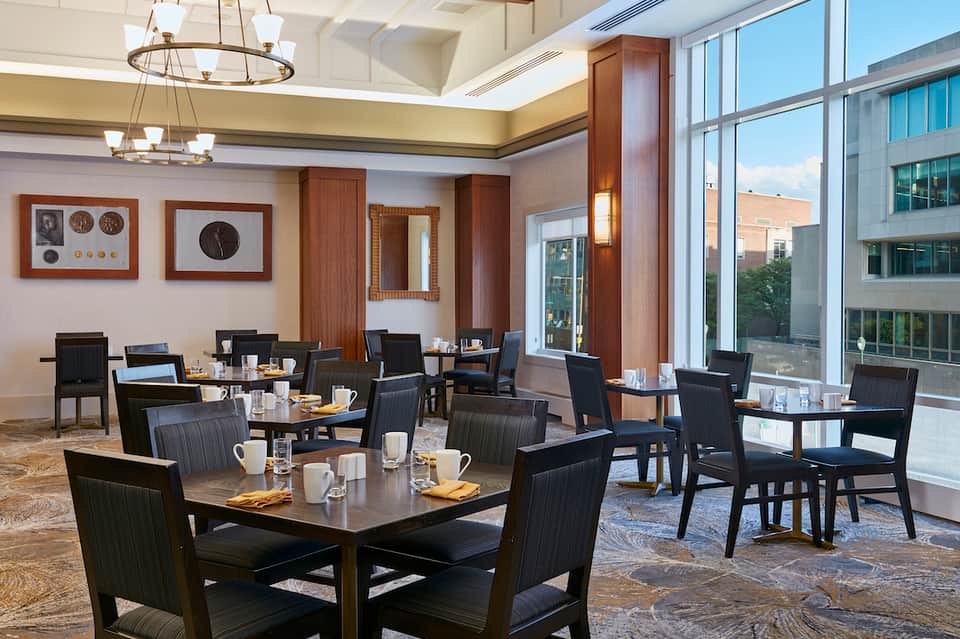 Modern breakfast room with black chairs, wooden tables, decorative wall clocks, and floor-to-ceiling windows with city views