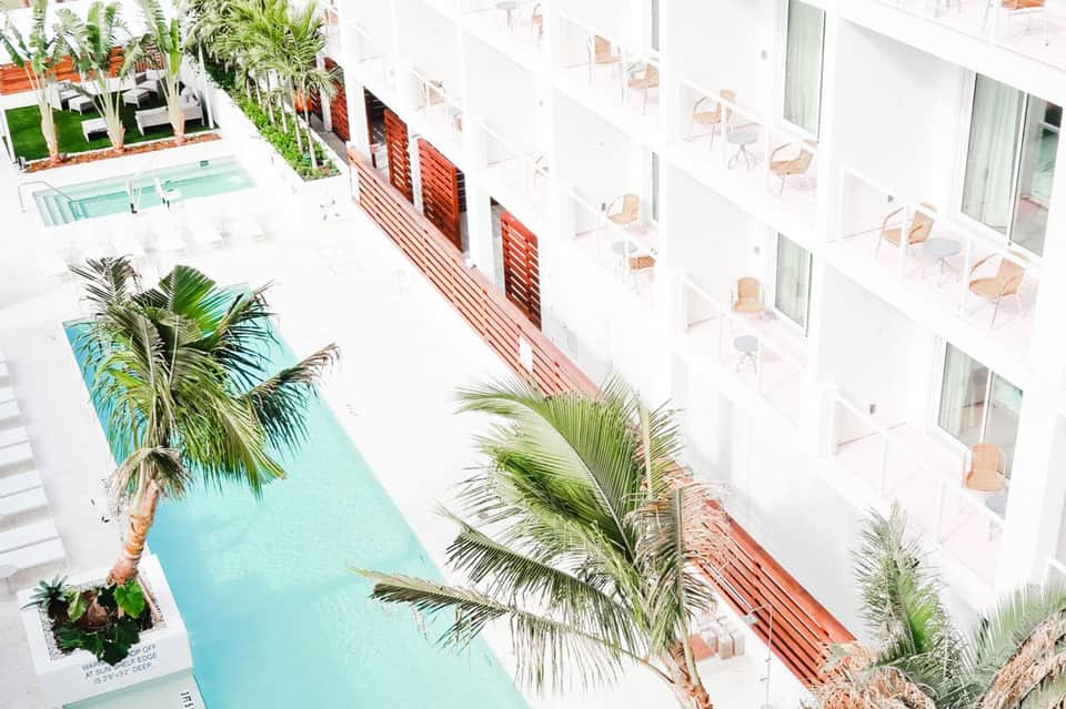 Overhead view of modern hotel courtyard with turquoise pool, palm trees, and white building