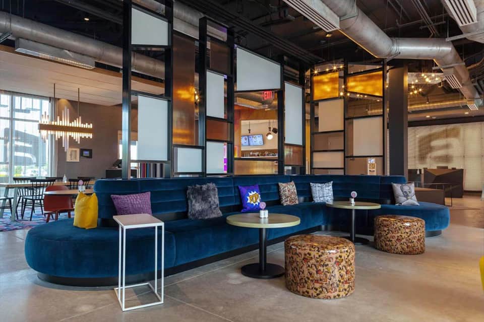 Modern hotel lobby with curved blue velvet sofa, patterned ottomans, pendant lights, and industrial ceiling design