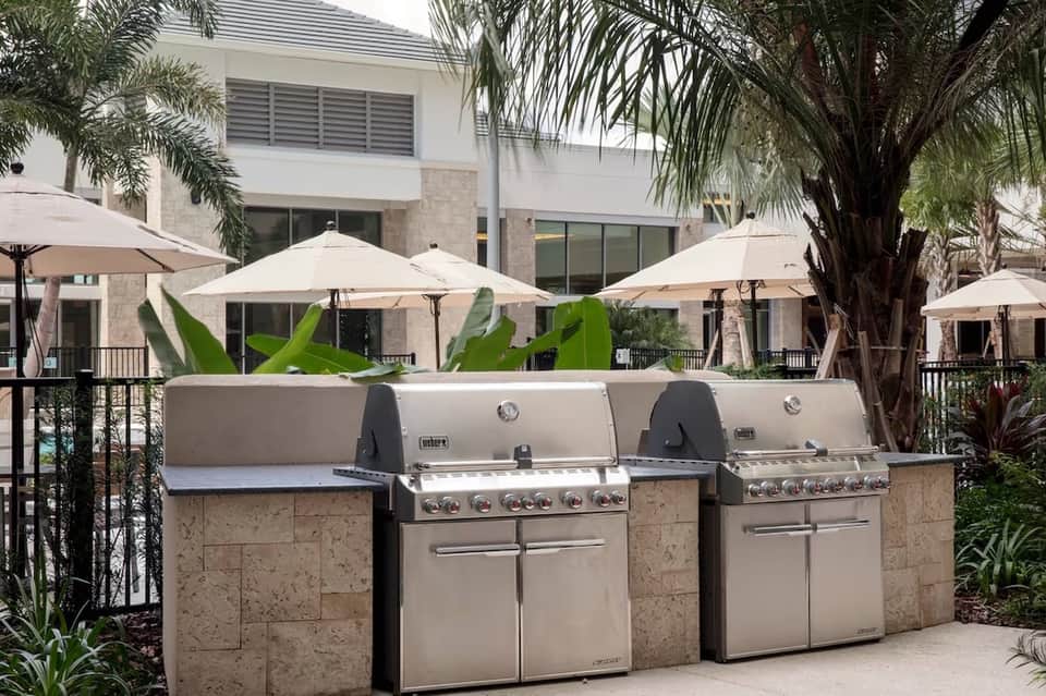 Outdoor patio area with three stainless steel grills, palm trees, and white umbrellas in front of resort building.