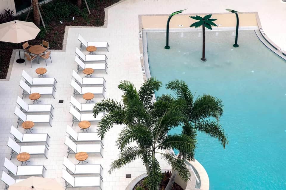 Aerial view of resort pool with turquoise water, loungers with tables, palm trees, and white patio area