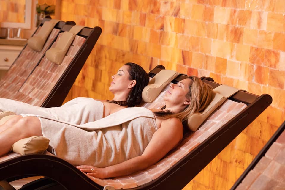 Two women relaxing on spa massage beds against an orange brick wall