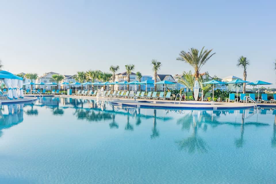 Resort pool area with turquoise water, palm trees, blue umbrellas, and waterfront buildings under clear sky
