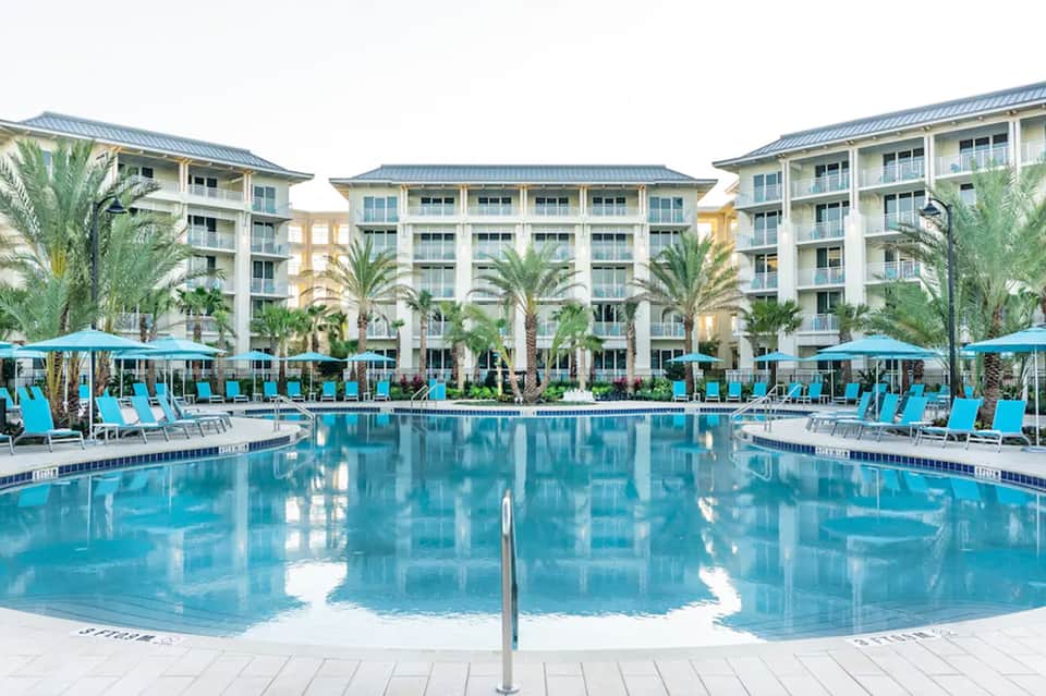 Large resort pool with bright turquoise water, palm trees, colorful lounge chairs, and multi-story hotel buildings