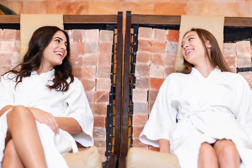 Two women in white robes sitting on spa chairs in front of brick wall, smiling at each other