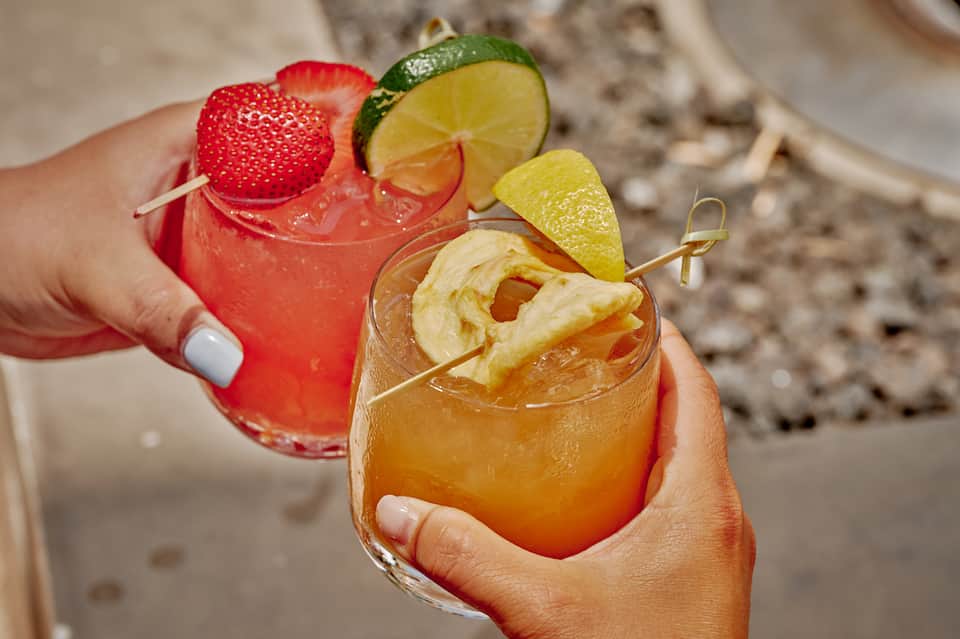 Two colorful cocktails with fruit garnishes held in hands at a beach setting