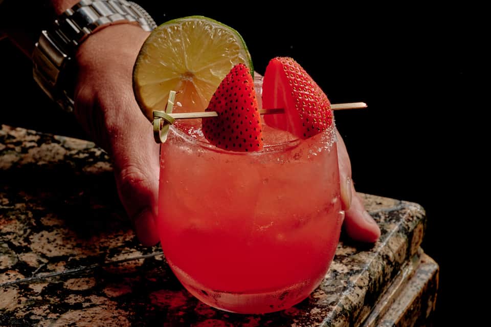 Strawberry cocktail with lime slice garnished on dark background, held by person wearing watch