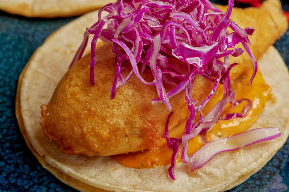 Gourmet taco on white plate topped with crispy fish and purple pickled onions