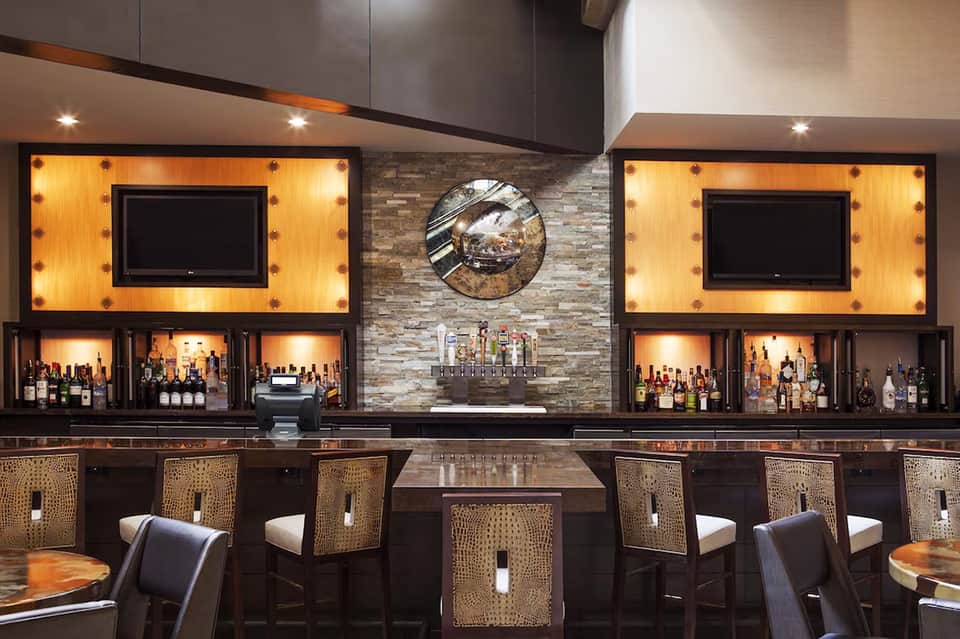 Sophisticated hotel bar with illuminated shelving, TVs, stone accent wall, and woven bar stools