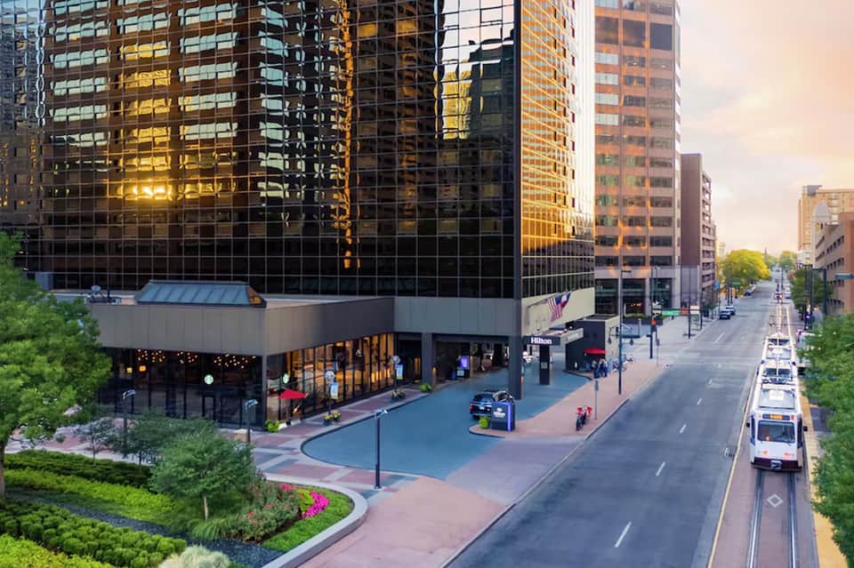 Urban hotel entrance at dusk with glass facade, illuminated ground plaza, and tall buildings on city street
