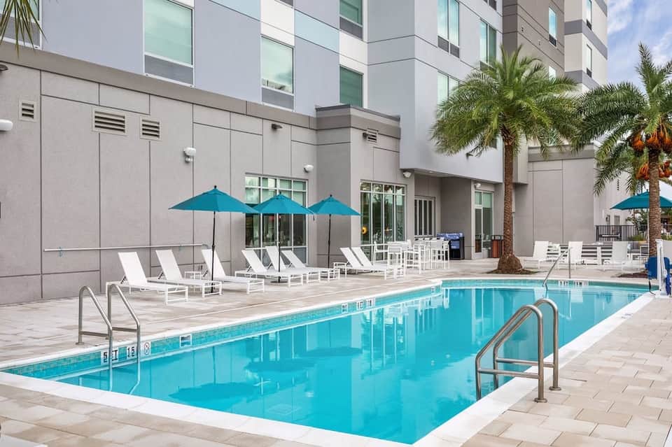Contemporary hotel pool deck with turquoise water, white lounge chairs, blue umbrellas, and modern building