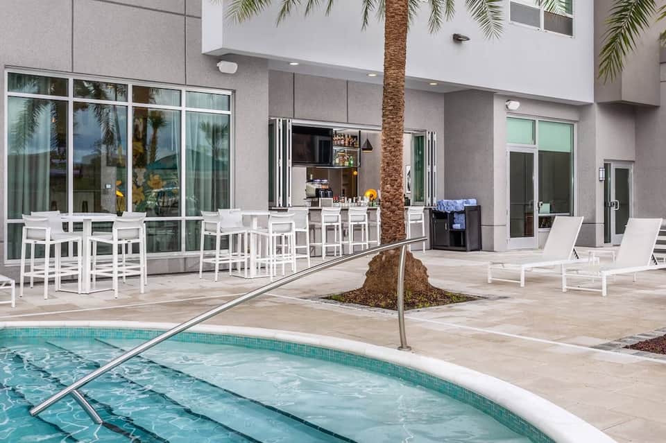 Modern poolside terrace with white bar seating, palm tree, and contemporary building architecture