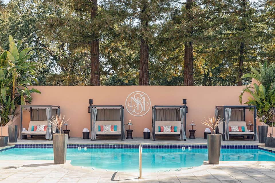 Luxury resort pool with cabanas and pink wall backdrop surrounded by tall trees at sunset