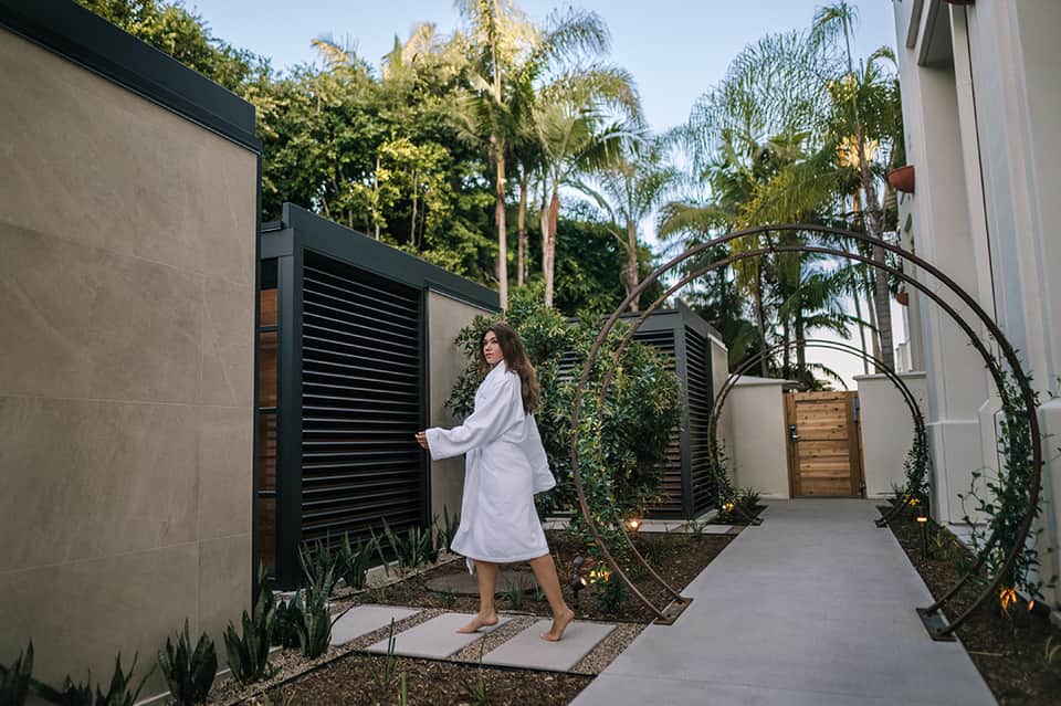 Woman in white robe walking through modern spa courtyard with black louvered gates
