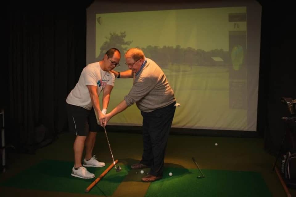 Two men playing indoor golf simulator with projected course display on screen