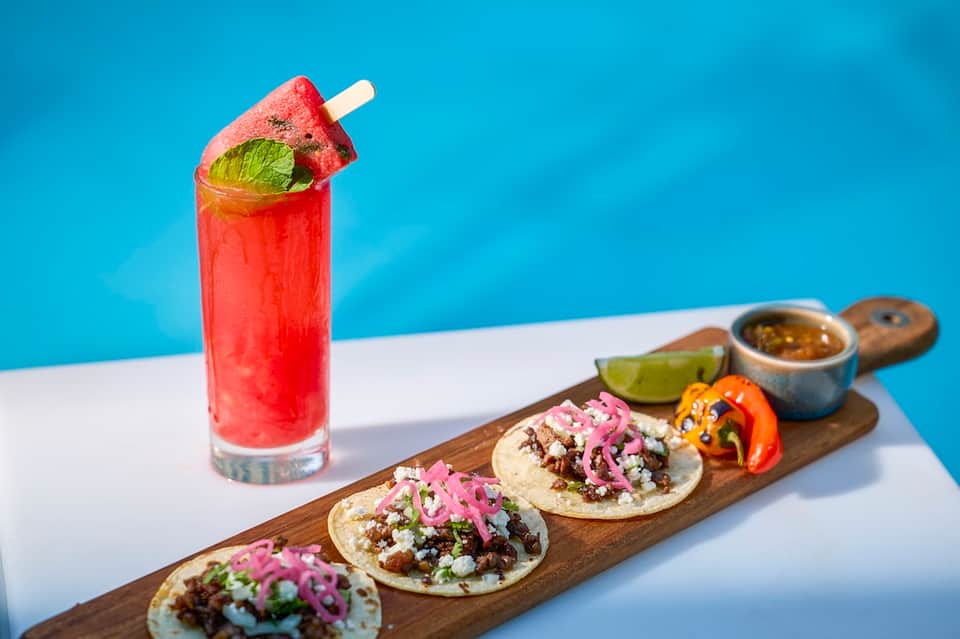 Tropical cocktail with watermelon popsicle, tacos with pickled onions, lime, and salsa on wooden board