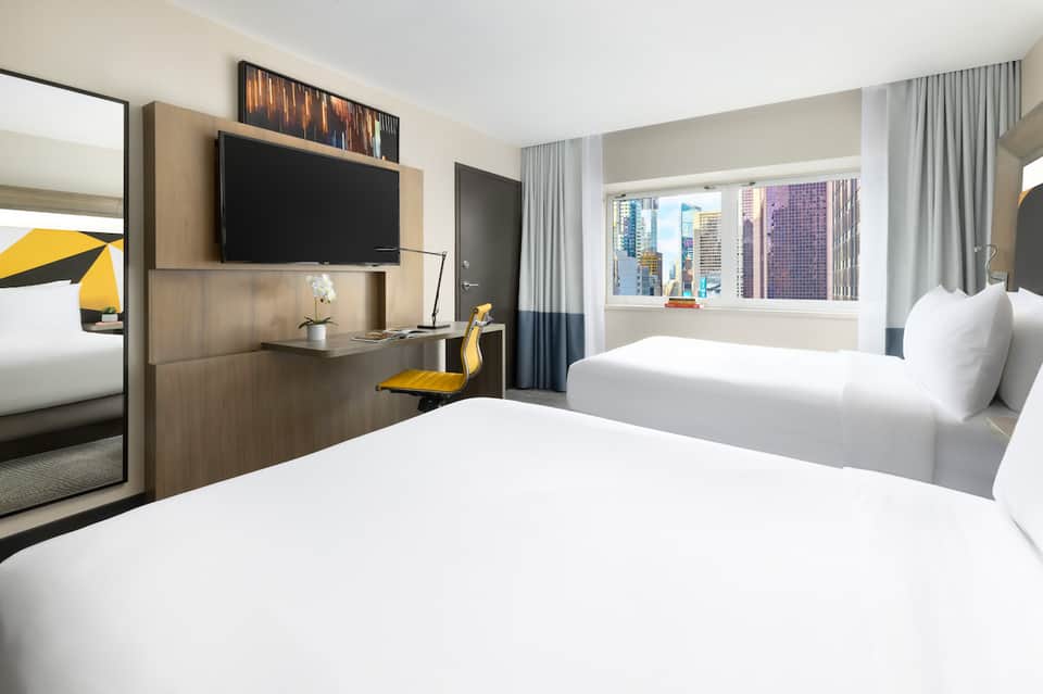 Contemporary hotel room with twin beds, flat-screen TV, wooden desk, and city skyline views through windows