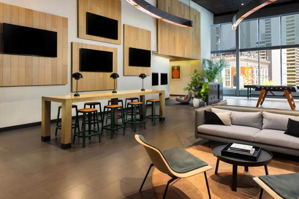 Contemporary hotel lounge with wooden accent wall, multiple TV screens, bar seating, sofa, and floor-to-ceiling windows