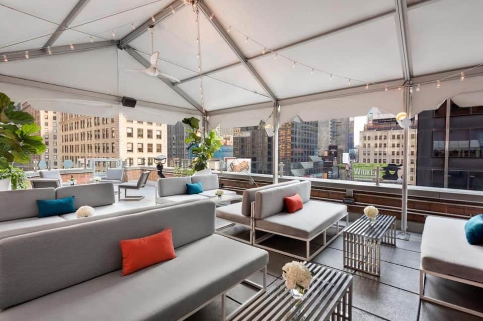 Modern rooftop lounge with gray seating, colorful pillows, city skyline views through glass walls, string lighting