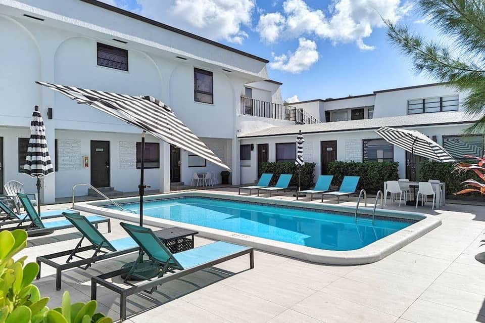 Resort courtyard with rectangular swimming pool, striped umbrellas, lounge chairs, and white modern architecture