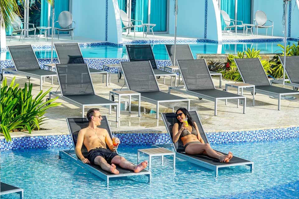 Two guests relaxing on lounge chairs in shallow pool with tropical setting and blue umbrellas