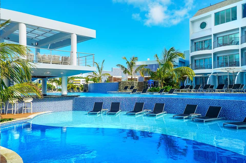 Modern resort pool surrounded by tropical palms, loungers, and multi-story blue-toned building with covered pavilion