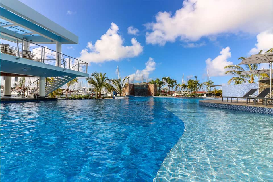 Resort swimming pool with palm trees, covered structures, and sailboats in background