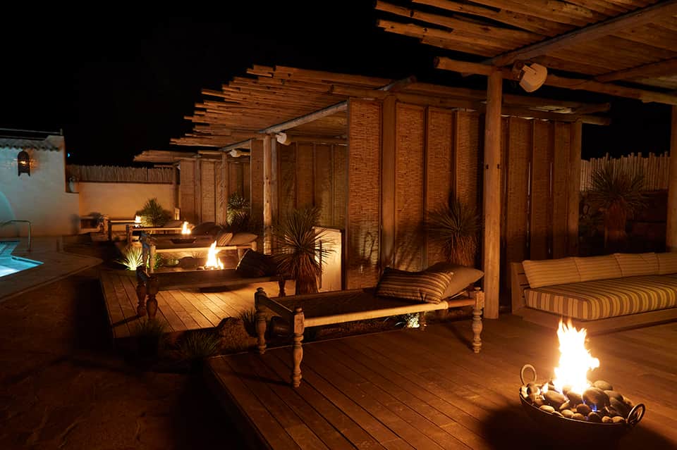 Nighttime rooftop terrace with fire pits, wooden decking, lounge seating, and brick privacy walls