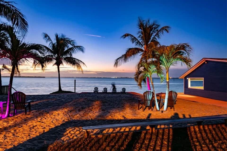Beach at dusk with palm trees, Adirondack chairs, surfboards, and illuminated beach house
