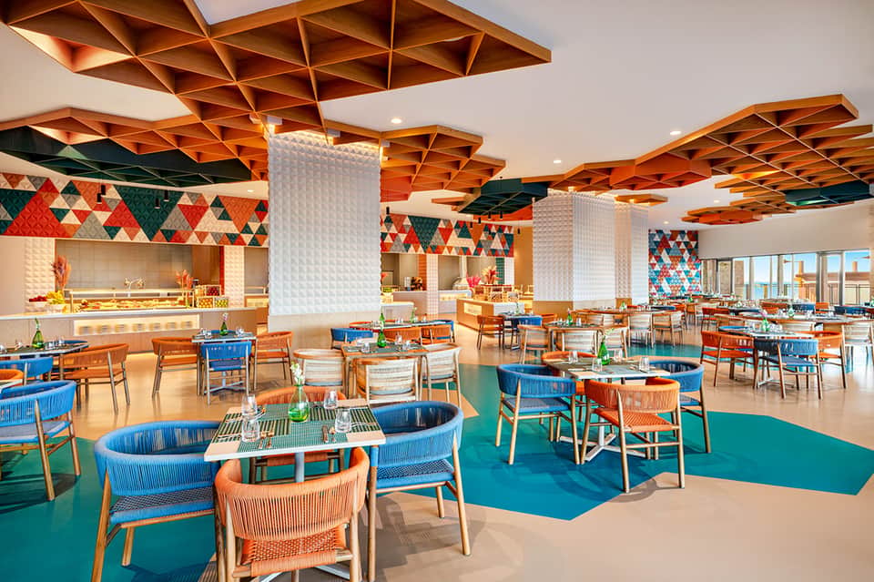 Bright dining room with geometric wooden ceiling panels, blue and orange furniture, and open kitchen