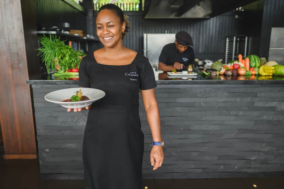 Chef presenting plated dish at modern open kitchen with fresh produce display